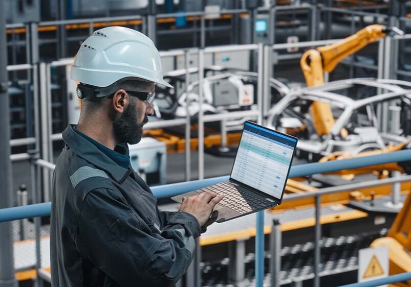 Car Factory Engineer in Work Uniform Using Laptop Computer with Spreadsheet Software. Working with Software at Automotive Industrial Manufacturing Facility Dedicated for Vehicle Production.