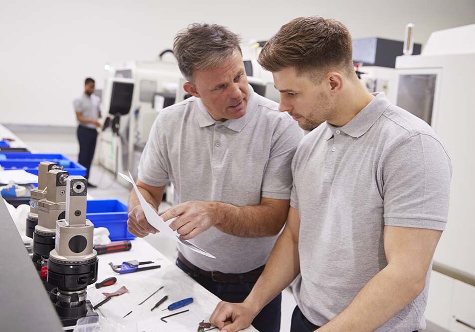Engineer And Apprentice Discussing Job Sheet In Factory